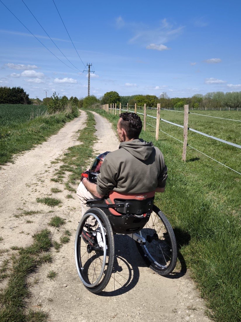 Axel Broutin dans son fauteuil tout-terrain sur un chemin de campagne, avançant sous un grand ciel bleu